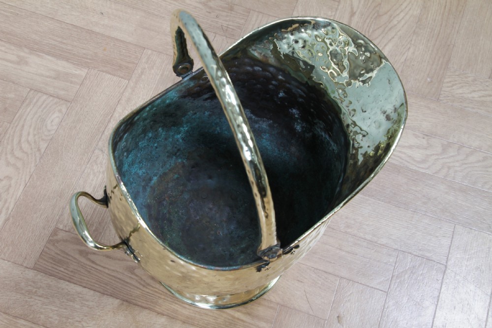 A Victorian hand-hammered brass helmet-shaped coal scuttle featuring a top swing handle, rear loop handle, and a round pedestal base. View IX