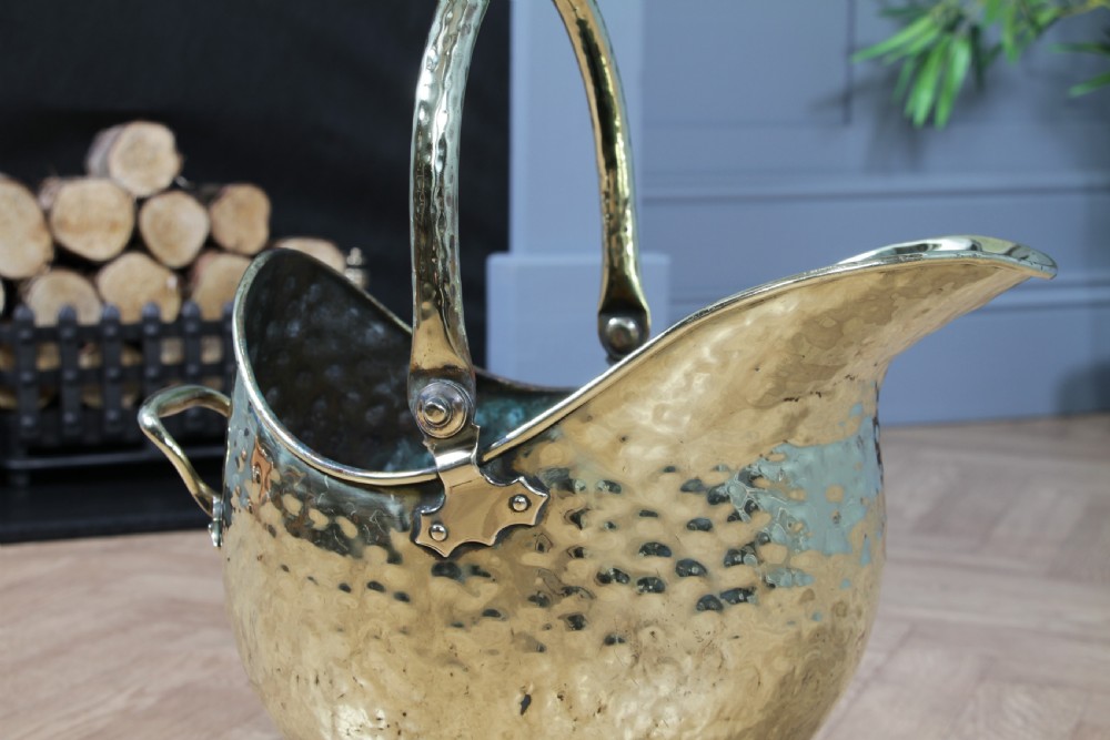 A Victorian hand-hammered brass helmet-shaped coal scuttle featuring a top swing handle, rear loop handle, and a round pedestal base. View X