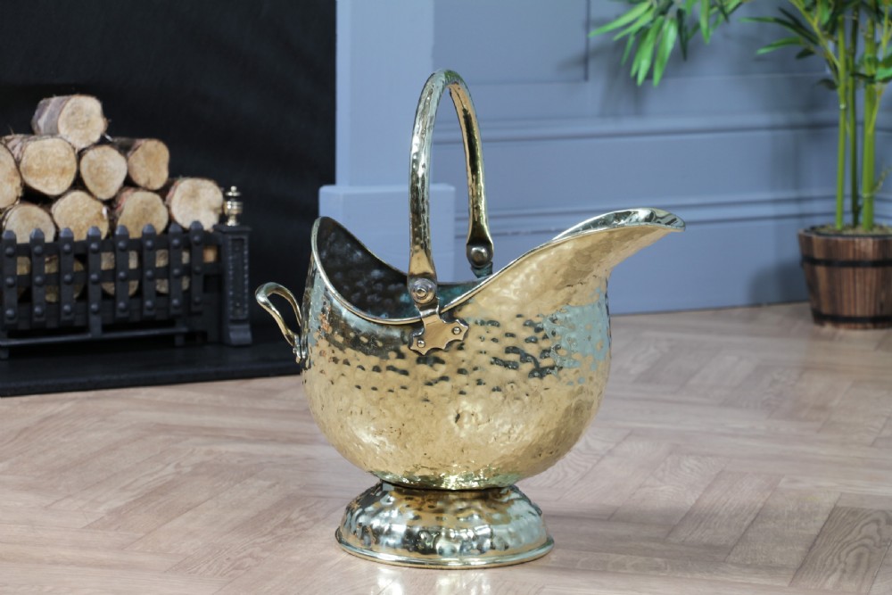 A Victorian hand-hammered brass helmet-shaped coal scuttle featuring a top swing handle, rear loop handle, and a round pedestal base. View XI