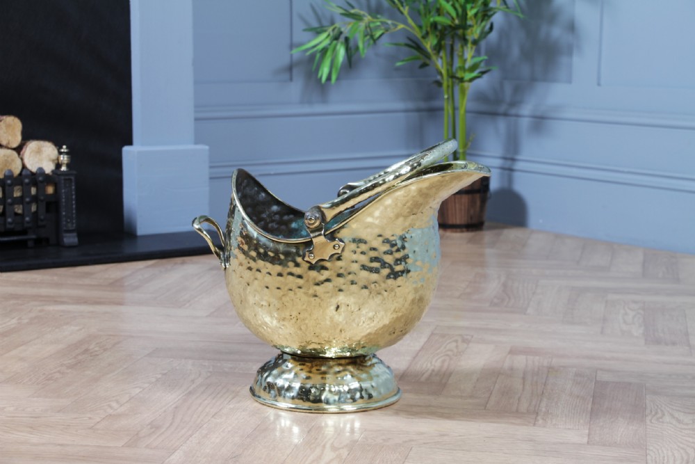 A Victorian hand-hammered brass helmet-shaped coal scuttle featuring a top swing handle, rear loop handle, and a round pedestal base. View XII