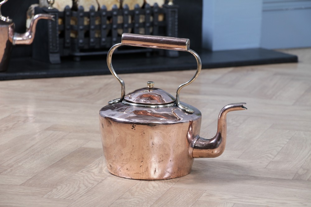 An antique George III polished copper kettle featuring a brass finial lid, overhead handle, and curved spout.