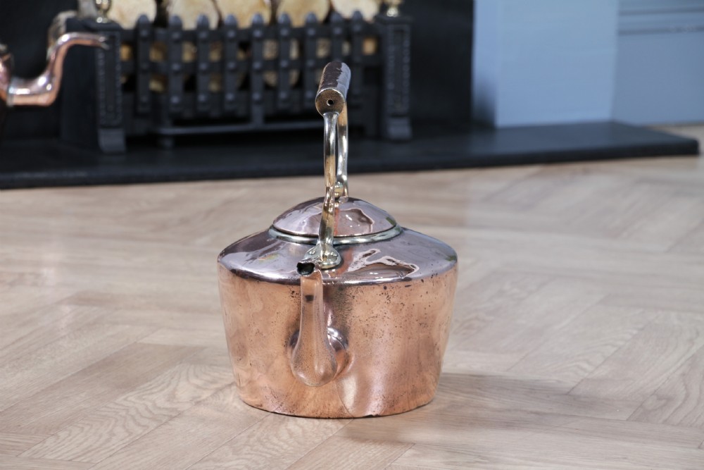 An antique George III polished copper kettle featuring a brass finial lid, overhead handle, and curved spout. View V