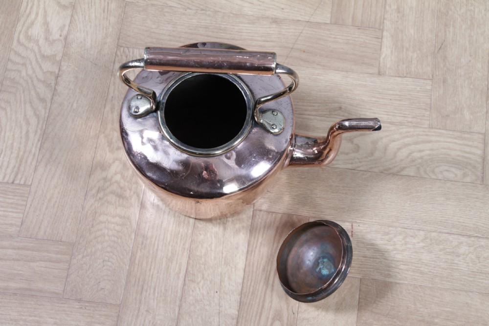 An antique George III polished copper kettle featuring a brass finial lid, overhead handle, and curved spout. View VII