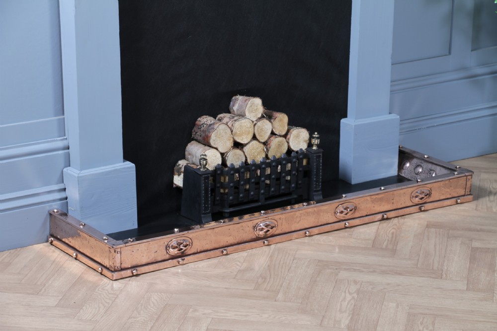 A low polished copper fireplace fender curb featuring hand-riveted edges and decorative embossed oval motifs around the perimeter.