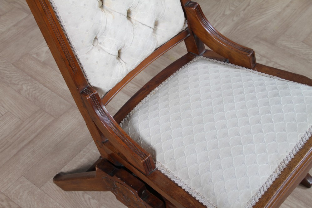 An Edwardian walnut platform rocking chair featuring cream button-backed upholstery and turned wooden spindles on a stationary crossed base. View X