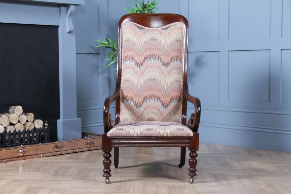 A high-backed Victorian mahogany fireside armchair featuring geometric flame-patterned upholstery, curved scroll armrests, turned front legs on brass castors, and a dark polished finish. View VI