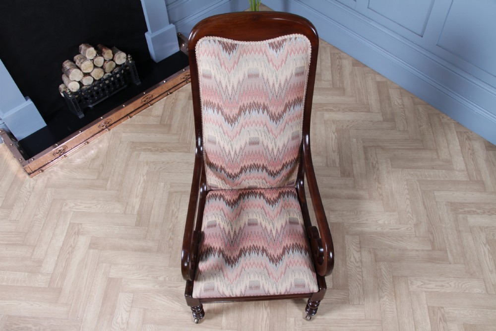 A high-backed Victorian mahogany fireside armchair featuring geometric flame-patterned upholstery, curved scroll armrests, turned front legs on brass castors, and a dark polished finish. View XV
