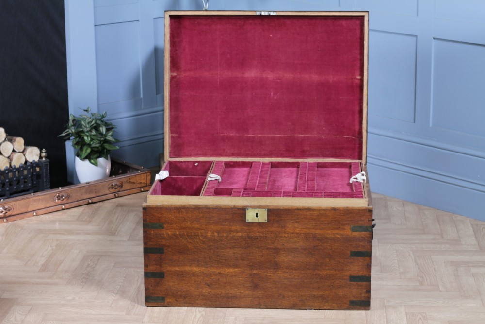 An open Victorian oak campaign trunk featuring brass-bound corners, a central brass keyhole escutcheon, and a fitted interior lined in deep red velvet.