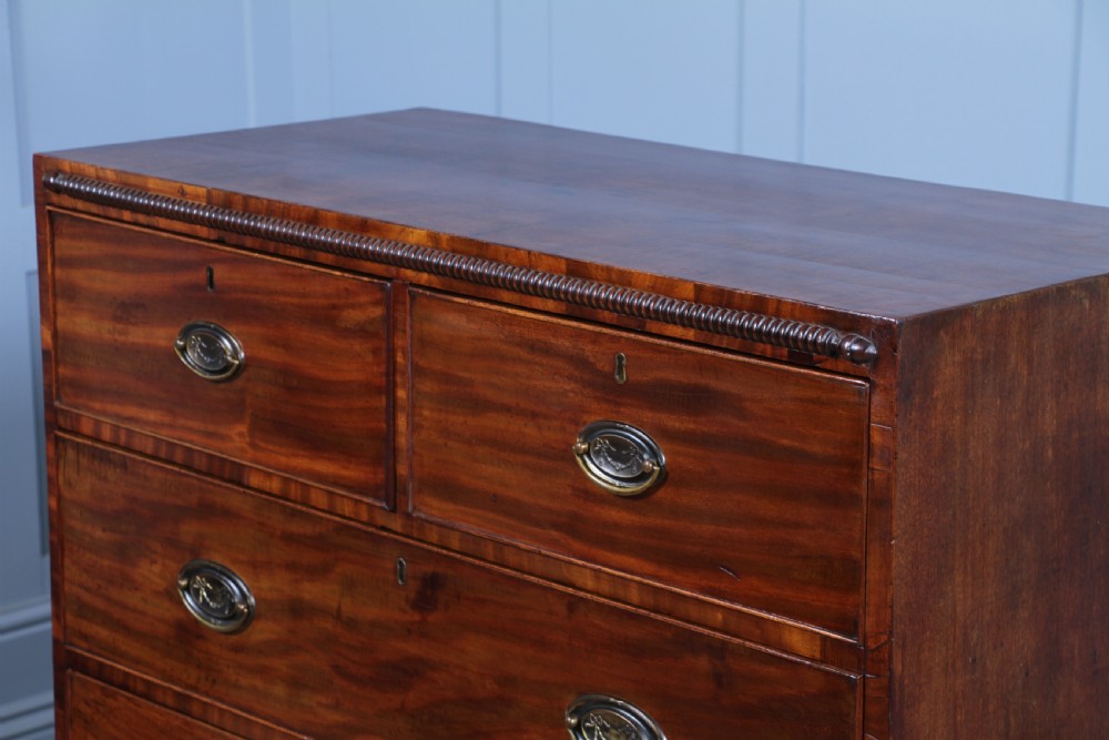 A William IV mahogany chest of drawers featuring two short over three long drawers with oval brass handles and turned feet. View II