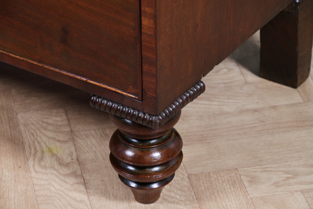 A William IV mahogany chest of drawers featuring two short over three long drawers with oval brass handles and turned feet. View X