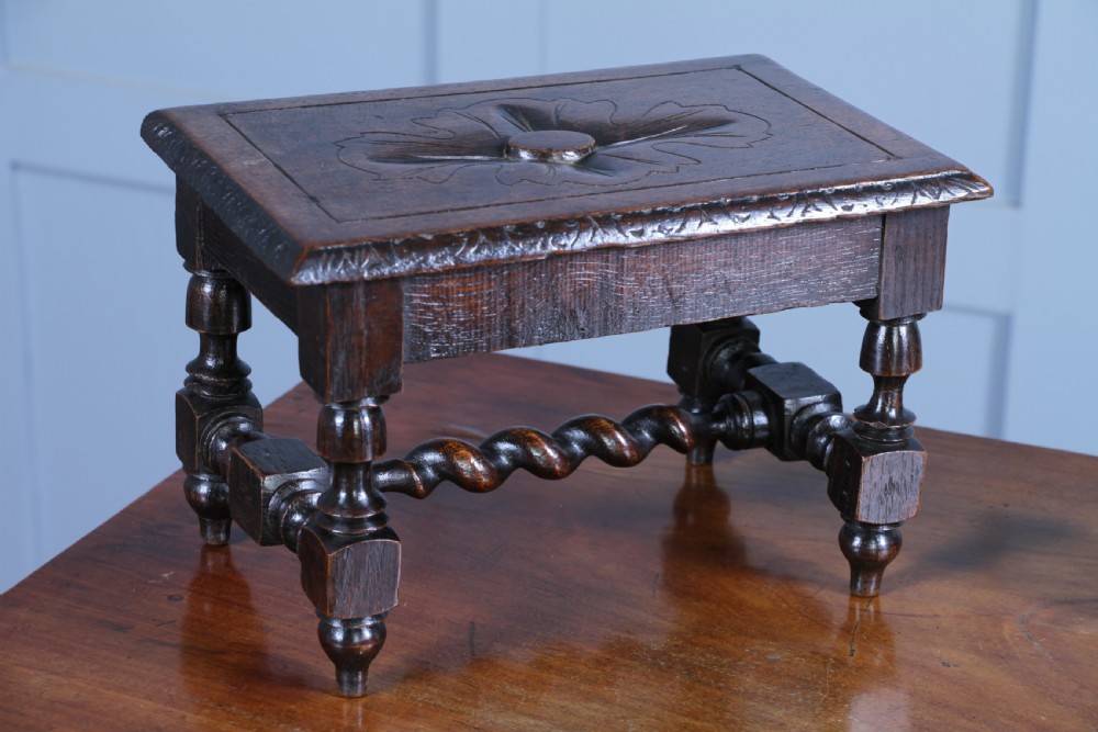 A small dark brown oak miniature stool with relief botanical carving on top, carved side aprons, turned legs, and a central barley twist stretcher.