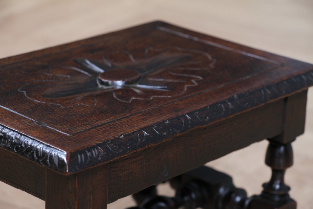 A small dark brown oak miniature stool with relief botanical carving on top, carved side aprons, turned legs, and a central barley twist stretcher. View VI
