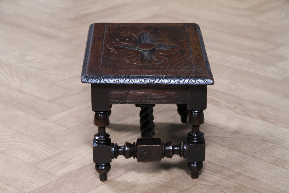 A small dark brown oak miniature stool with relief botanical carving on top, carved side aprons, turned legs, and a central barley twist stretcher. View VII