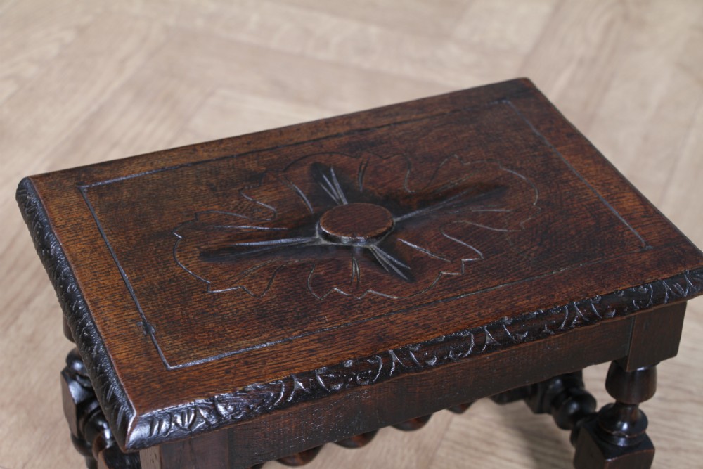 A small dark brown oak miniature stool with relief botanical carving on top, carved side aprons, turned legs, and a central barley twist stretcher. View X