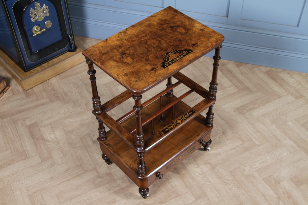 A Victorian burr walnut Canterbury whatnot featuring a flat top surface, turned supports, vertical dividers, a bottom drawer with inlaid motifs, and brass castors. View II