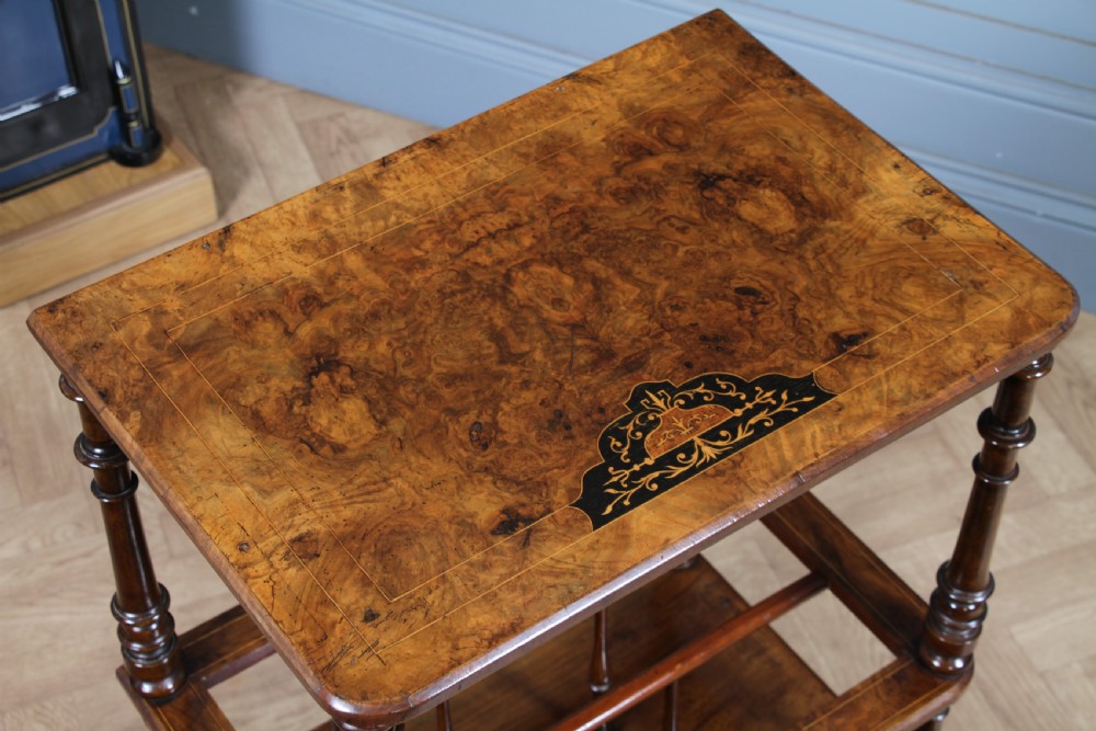 A Victorian burr walnut Canterbury whatnot featuring a flat top surface, turned supports, vertical dividers, a bottom drawer with inlaid motifs, and brass castors. View III