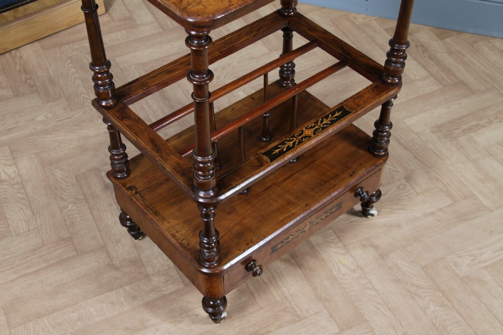 A Victorian burr walnut Canterbury whatnot featuring a flat top surface, turned supports, vertical dividers, a bottom drawer with inlaid motifs, and brass castors. View V