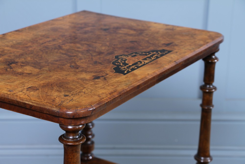 A Victorian burr walnut Canterbury whatnot featuring a flat top surface, turned supports, vertical dividers, a bottom drawer with inlaid motifs, and brass castors. View VI
