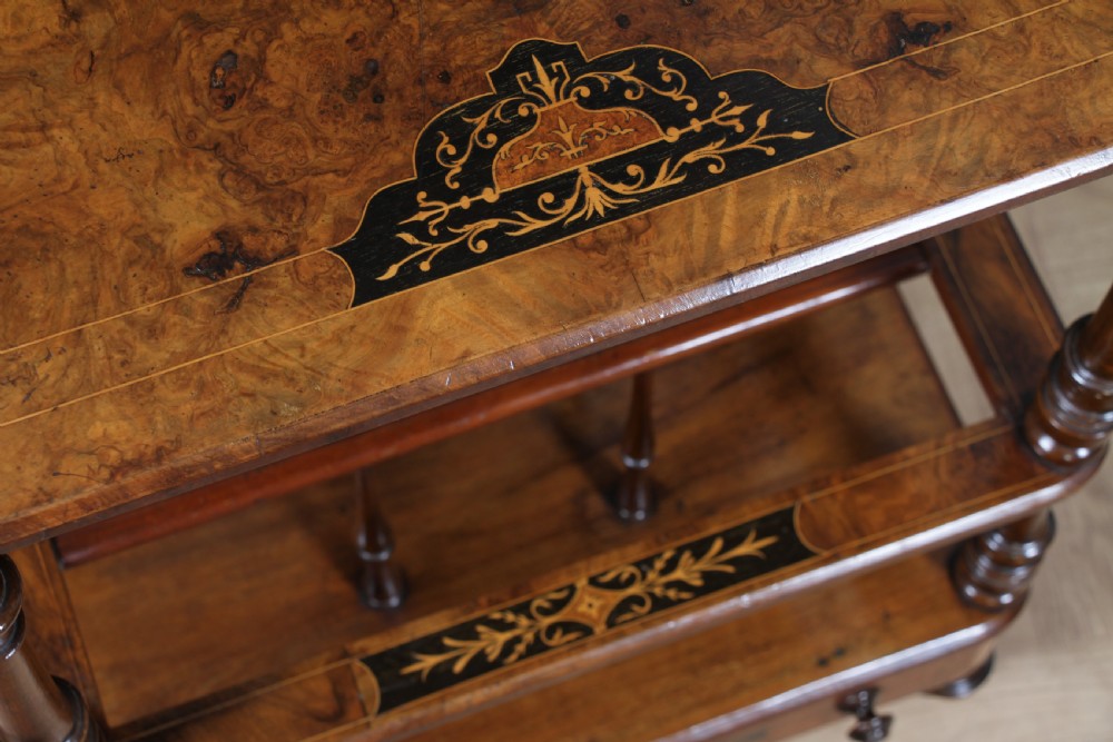 A Victorian burr walnut Canterbury whatnot featuring a flat top surface, turned supports, vertical dividers, a bottom drawer with inlaid motifs, and brass castors. View VII