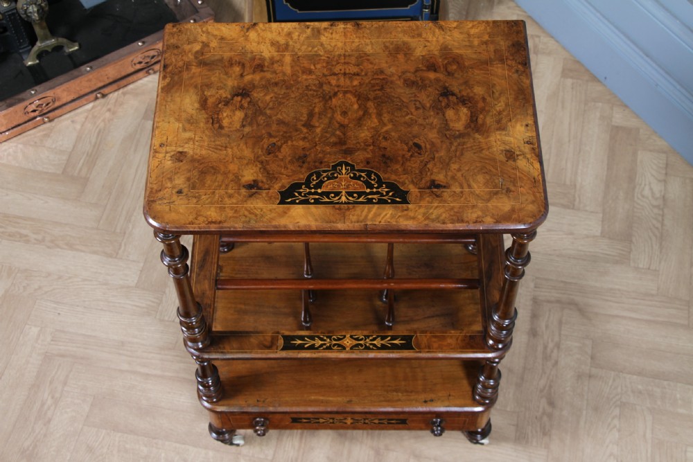 A Victorian burr walnut Canterbury whatnot featuring a flat top surface, turned supports, vertical dividers, a bottom drawer with inlaid motifs, and brass castors. View VIII