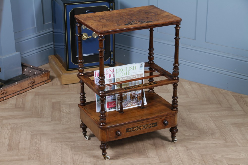 A Victorian burr walnut Canterbury whatnot featuring a flat top surface, turned supports, vertical dividers, a bottom drawer with inlaid motifs, and brass castors. View IX