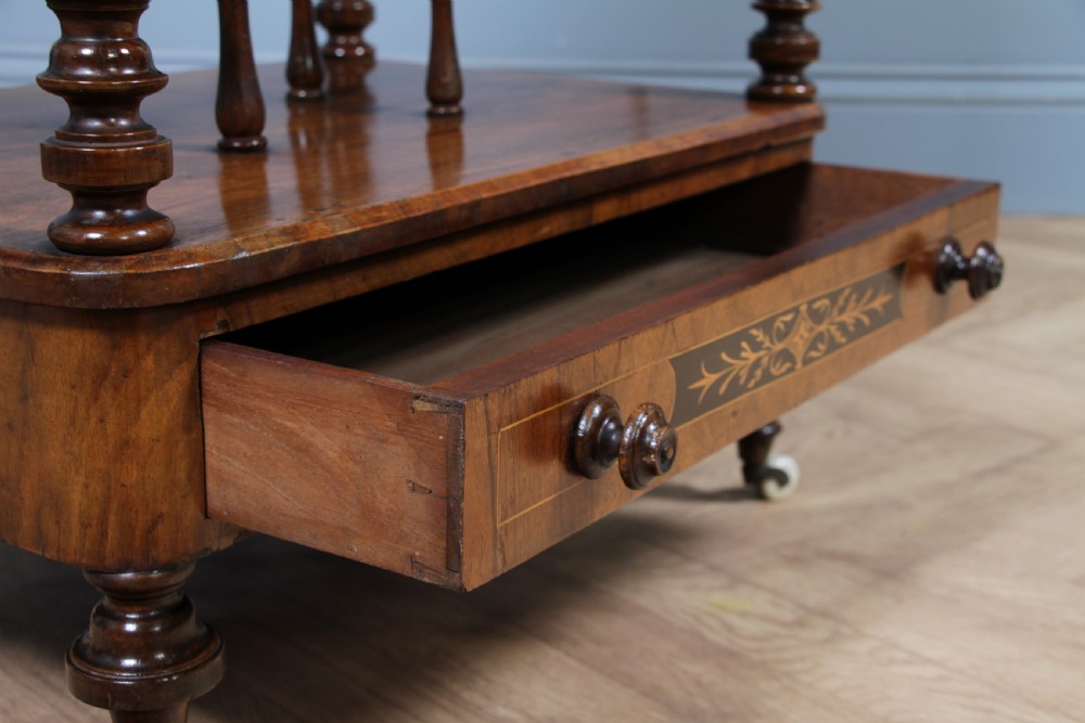 A Victorian burr walnut Canterbury whatnot featuring a flat top surface, turned supports, vertical dividers, a bottom drawer with inlaid motifs, and brass castors. View X