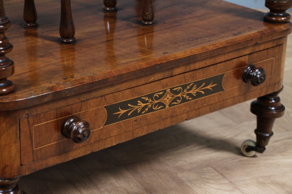 A Victorian burr walnut Canterbury whatnot featuring a flat top surface, turned supports, vertical dividers, a bottom drawer with inlaid motifs, and brass castors. View XI