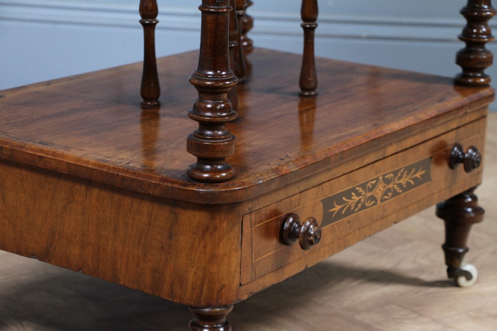 A Victorian burr walnut Canterbury whatnot featuring a flat top surface, turned supports, vertical dividers, a bottom drawer with inlaid motifs, and brass castors. View XII