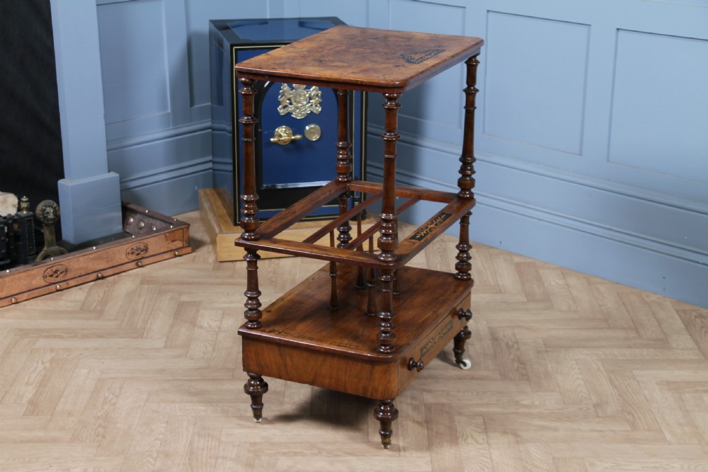 A Victorian burr walnut Canterbury whatnot featuring a flat top surface, turned supports, vertical dividers, a bottom drawer with inlaid motifs, and brass castors. View XV
