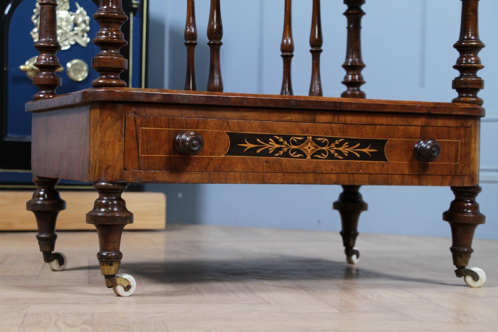 A Victorian burr walnut Canterbury whatnot featuring a flat top surface, turned supports, vertical dividers, a bottom drawer with inlaid motifs, and brass castors. View XVI