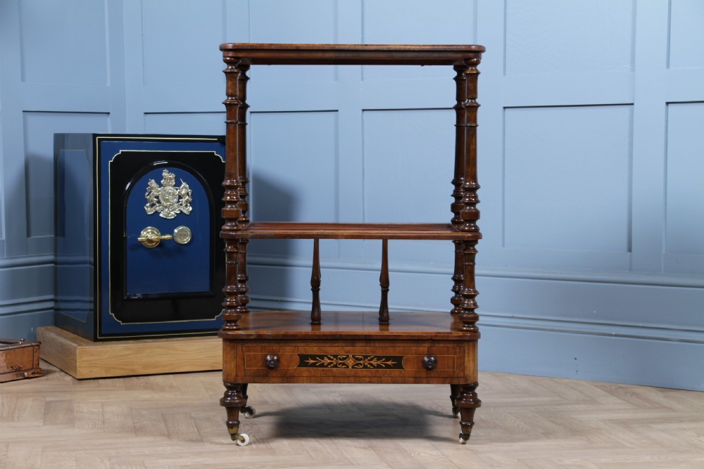 A Victorian burr walnut Canterbury whatnot featuring a flat top surface, turned supports, vertical dividers, a bottom drawer with inlaid motifs, and brass castors. View XVII