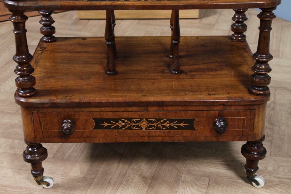 A Victorian burr walnut Canterbury whatnot featuring a flat top surface, turned supports, vertical dividers, a bottom drawer with inlaid motifs, and brass castors. View XVIII