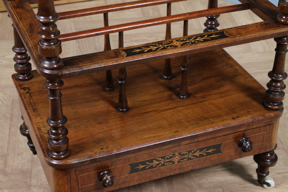 A Victorian burr walnut Canterbury whatnot featuring a flat top surface, turned supports, vertical dividers, a bottom drawer with inlaid motifs, and brass castors. View XIX