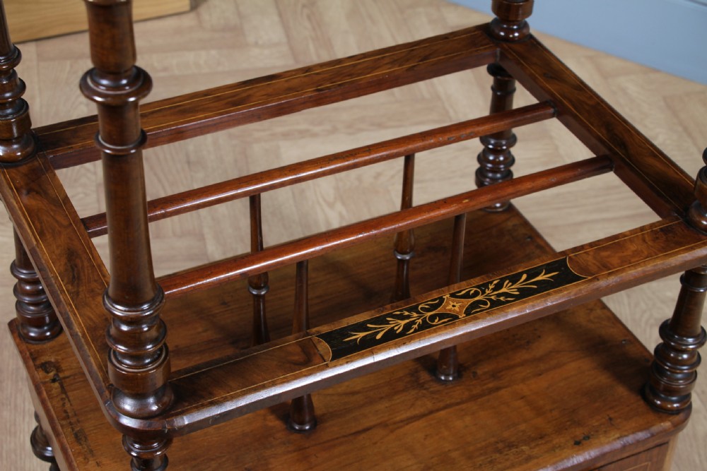 A Victorian burr walnut Canterbury whatnot featuring a flat top surface, turned supports, vertical dividers, a bottom drawer with inlaid motifs, and brass castors. View XX