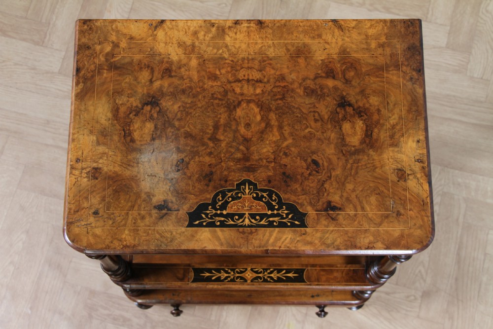 A Victorian burr walnut Canterbury whatnot featuring a flat top surface, turned supports, vertical dividers, a bottom drawer with inlaid motifs, and brass castors. View XXI