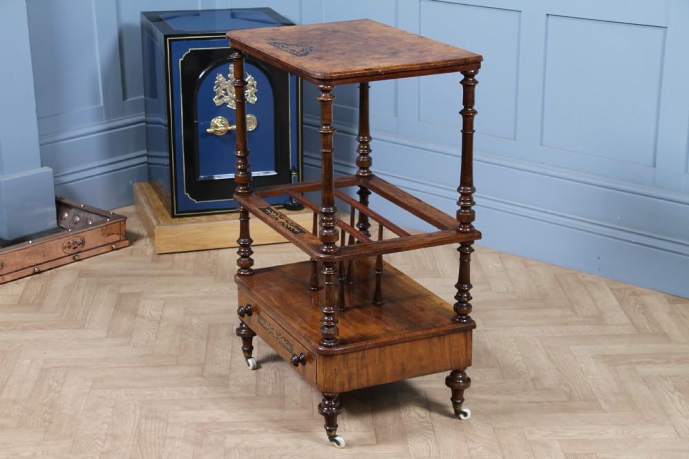 A Victorian burr walnut Canterbury whatnot featuring a flat top surface, turned supports, vertical dividers, a bottom drawer with inlaid motifs, and brass castors. View XXII