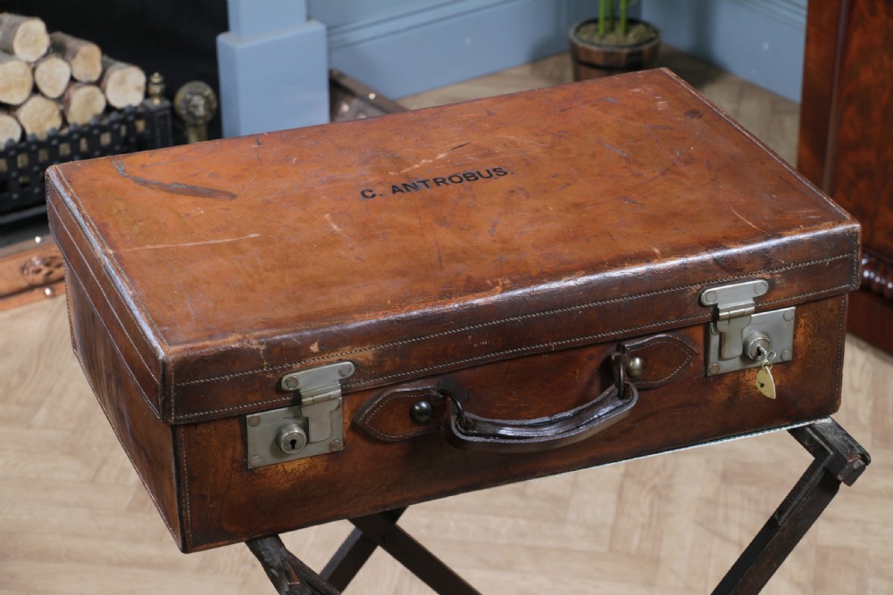 A rectangular chestnut brown leather suitcase with brass slide latches, a central leather handle, and black stencilled lettering reading C. ANTROBUS.