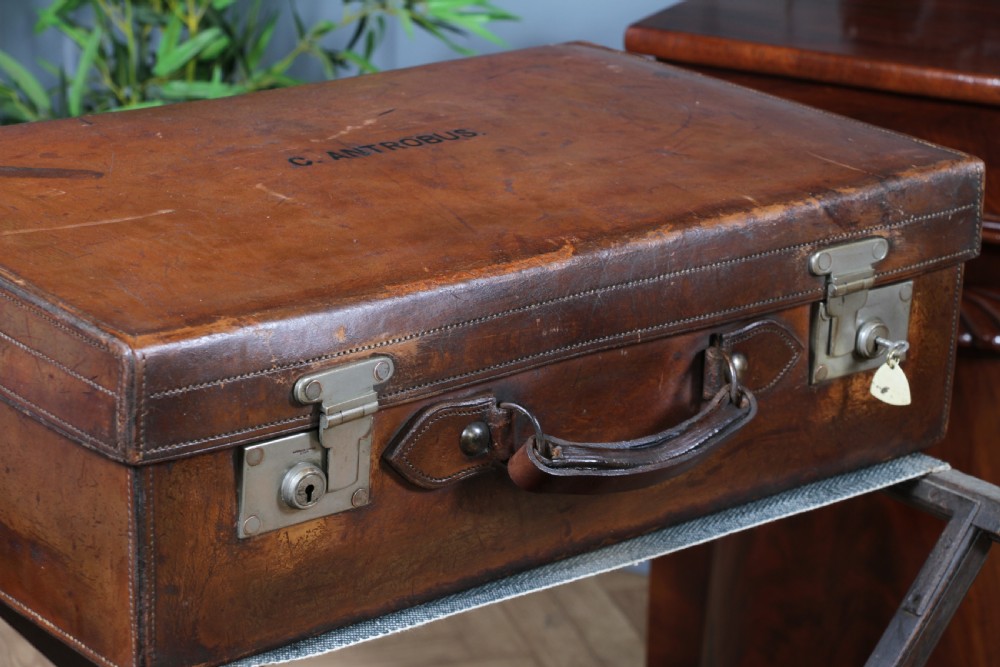 A rectangular chestnut brown leather suitcase with brass slide latches, a central leather handle, and black stencilled lettering reading C. ANTROBUS. View V