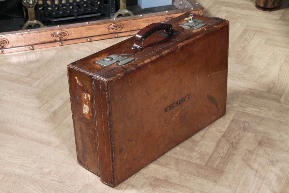 A rectangular chestnut brown leather suitcase with brass slide latches, a central leather handle, and black stencilled lettering reading C. ANTROBUS. View VII