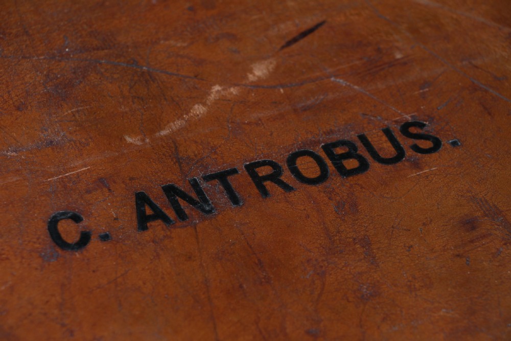 A rectangular chestnut brown leather suitcase with brass slide latches, a central leather handle, and black stencilled lettering reading C. ANTROBUS. View X