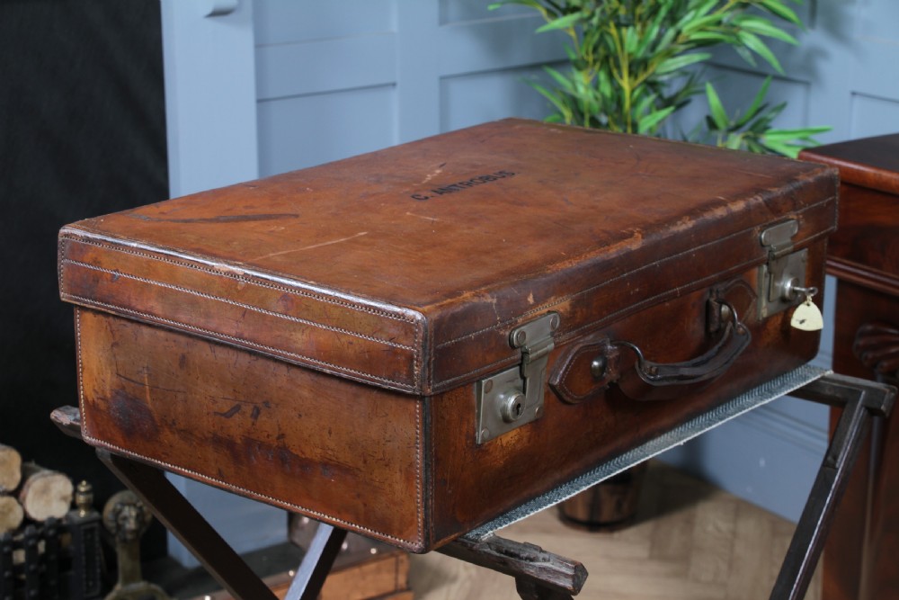 A rectangular chestnut brown leather suitcase with brass slide latches, a central leather handle, and black stencilled lettering reading C. ANTROBUS. View XV