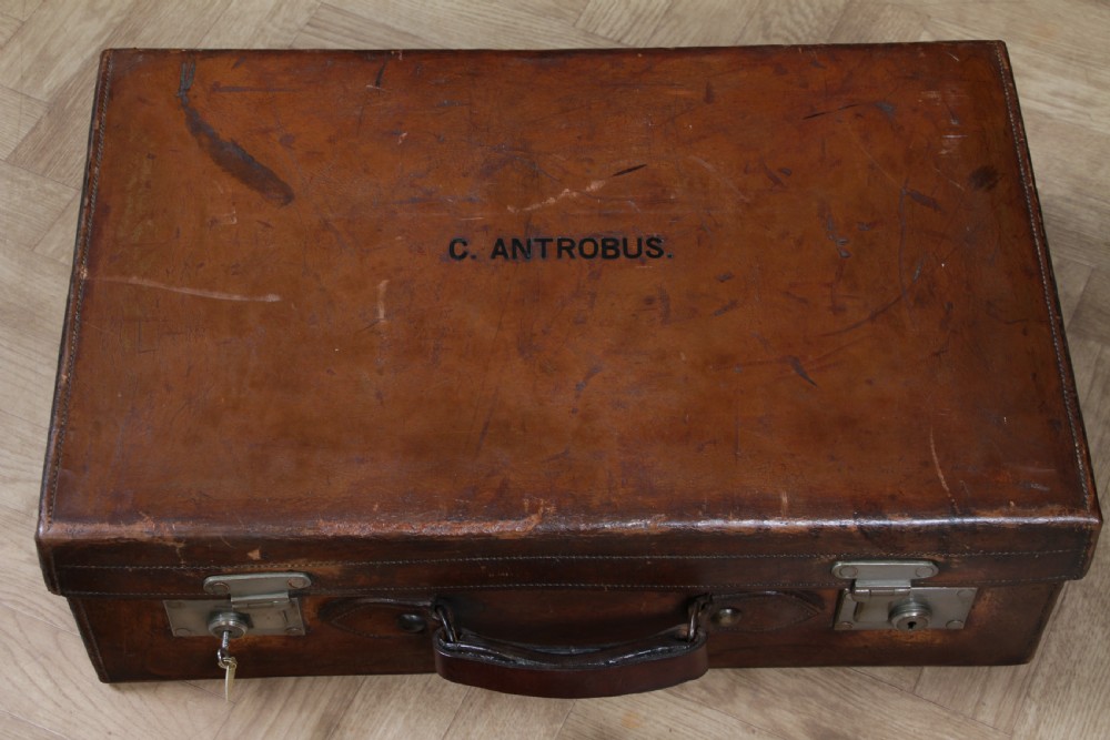 A rectangular chestnut brown leather suitcase with brass slide latches, a central leather handle, and black stencilled lettering reading C. ANTROBUS. View XXIII
