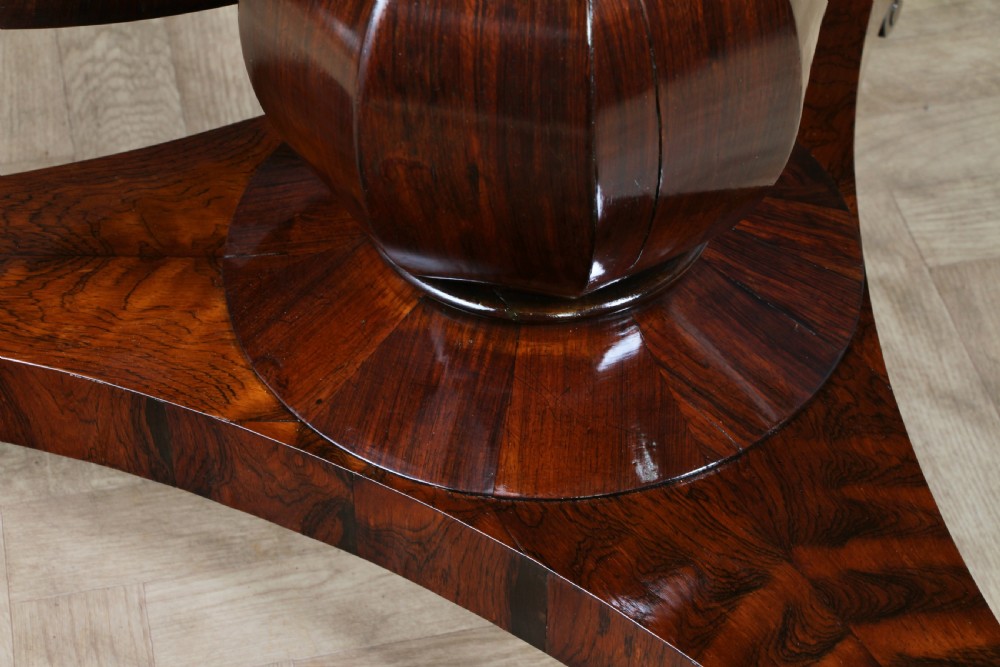 A circular dark rosewood centre table featuring a polished surface, a central bulbous faceted pedestal, and a triform base resting on carved scroll feet. View X