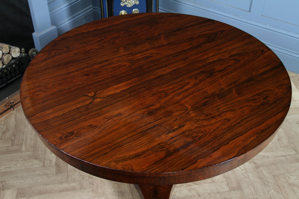 A circular dark rosewood centre table featuring a polished surface, a central bulbous faceted pedestal, and a triform base resting on carved scroll feet. View XXI