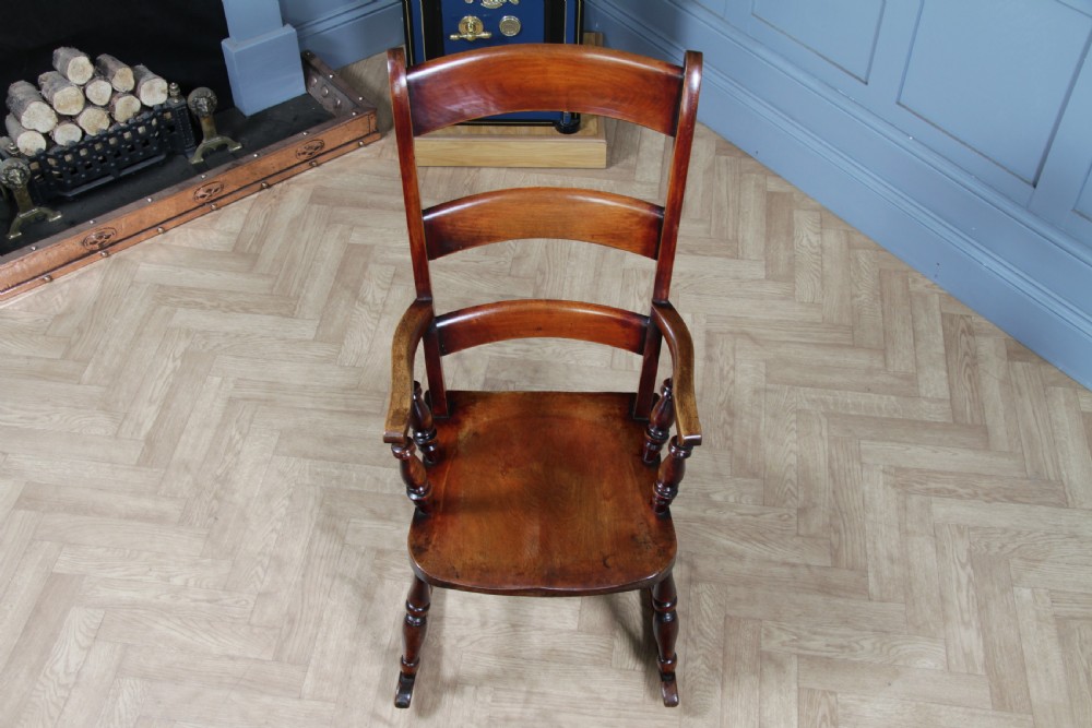 An antique Victorian ladder-back wooden rocking chair with three horizontal back slats, turned spindle arm supports, and curved rockers. View VI