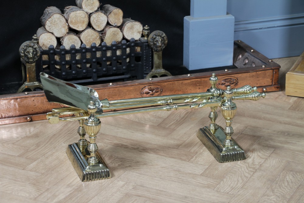 A late Victorian polished brass companion set featuring a shovel, tongs, and poker supported horizontally across two urn-topped brass firedogs. View II