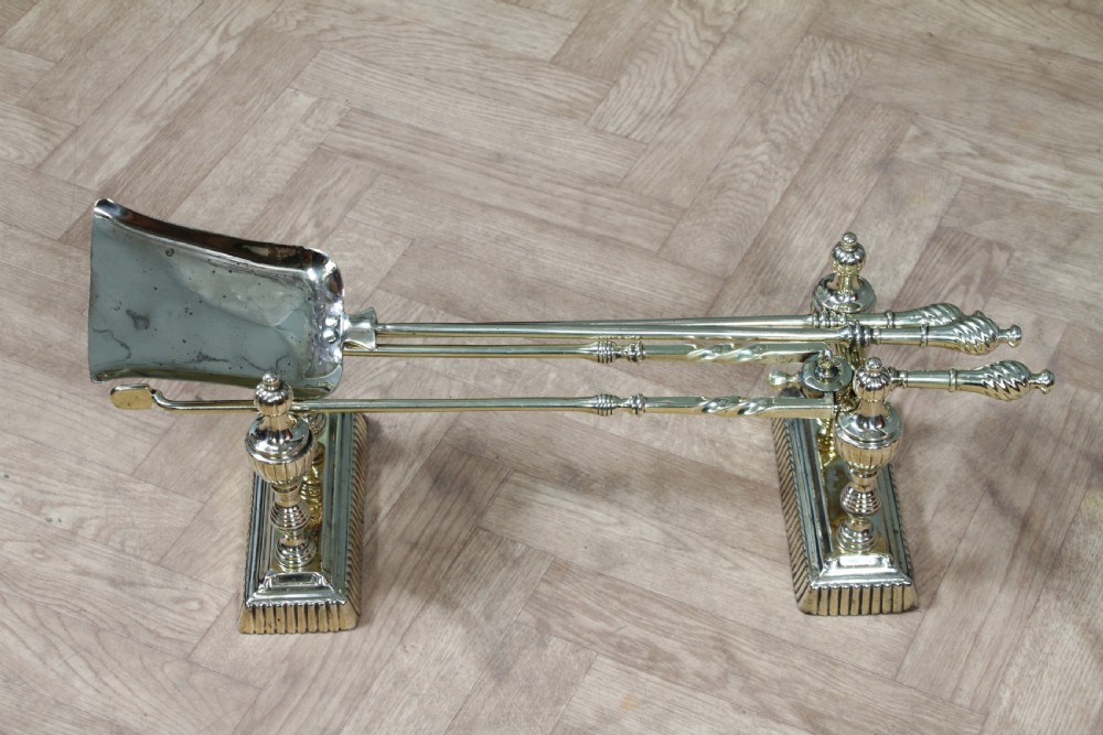 A late Victorian polished brass companion set featuring a shovel, tongs, and poker supported horizontally across two urn-topped brass firedogs. View III