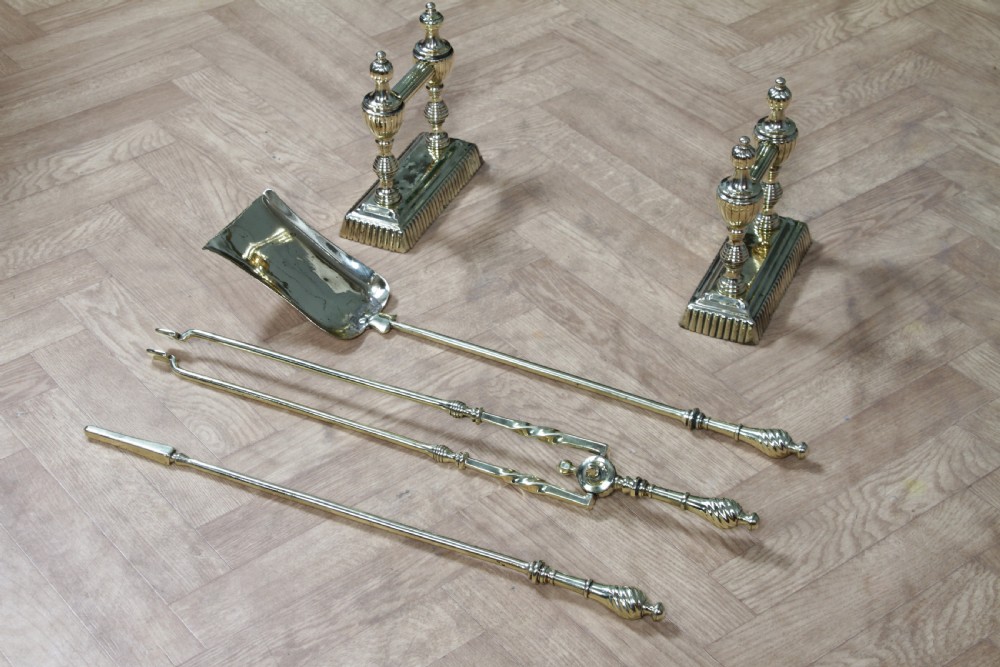 A late Victorian polished brass companion set featuring a shovel, tongs, and poker supported horizontally across two urn-topped brass firedogs. View V