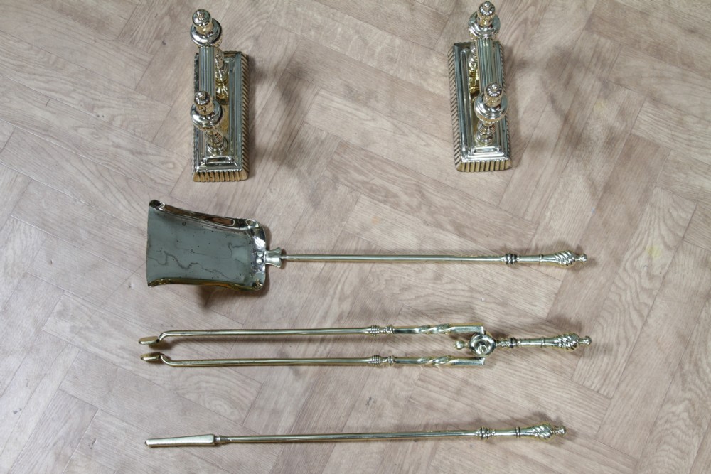A late Victorian polished brass companion set featuring a shovel, tongs, and poker supported horizontally across two urn-topped brass firedogs. View VII