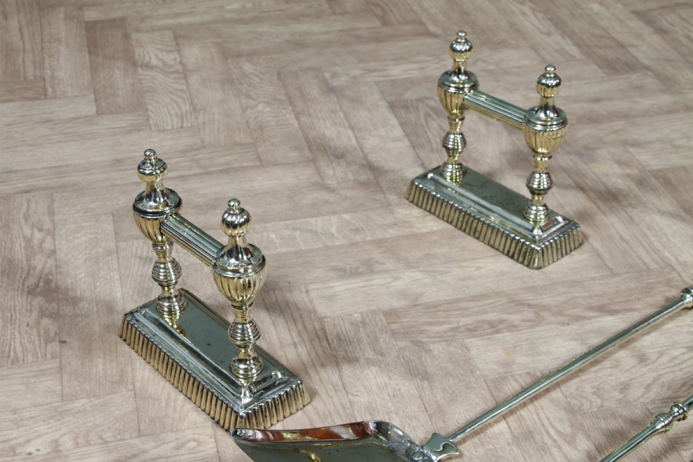 A late Victorian polished brass companion set featuring a shovel, tongs, and poker supported horizontally across two urn-topped brass firedogs. View VIII
